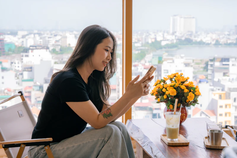 A young woman sitting by a big window, smiling at her phone.