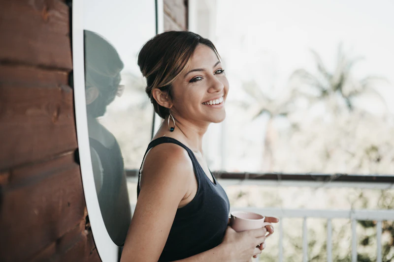 A woman smiling by a window in morning light, holding a mug.